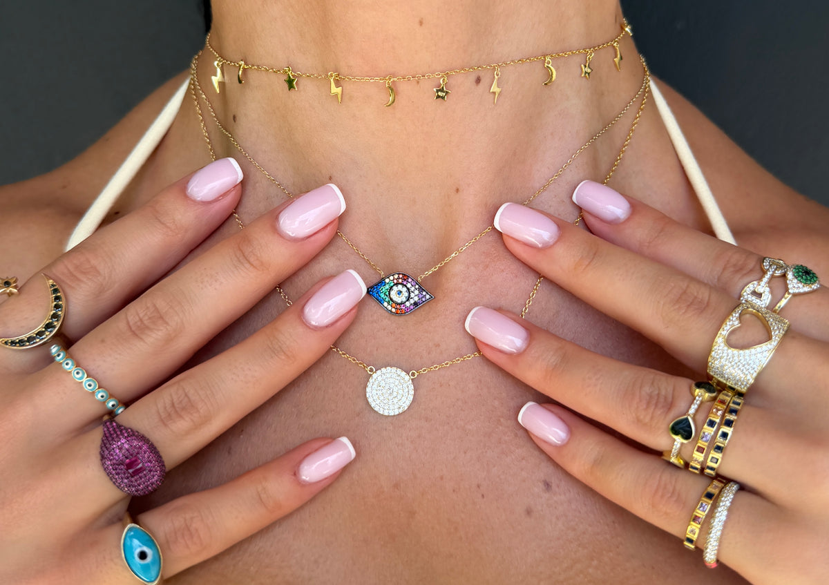 A close-up lifestyle shot of a model wearing multiple delicate gold necklaces, including a star-patterned choker, a blue and white evil eye pendant, and a silver disc charm, along with several statement rings on her hands.